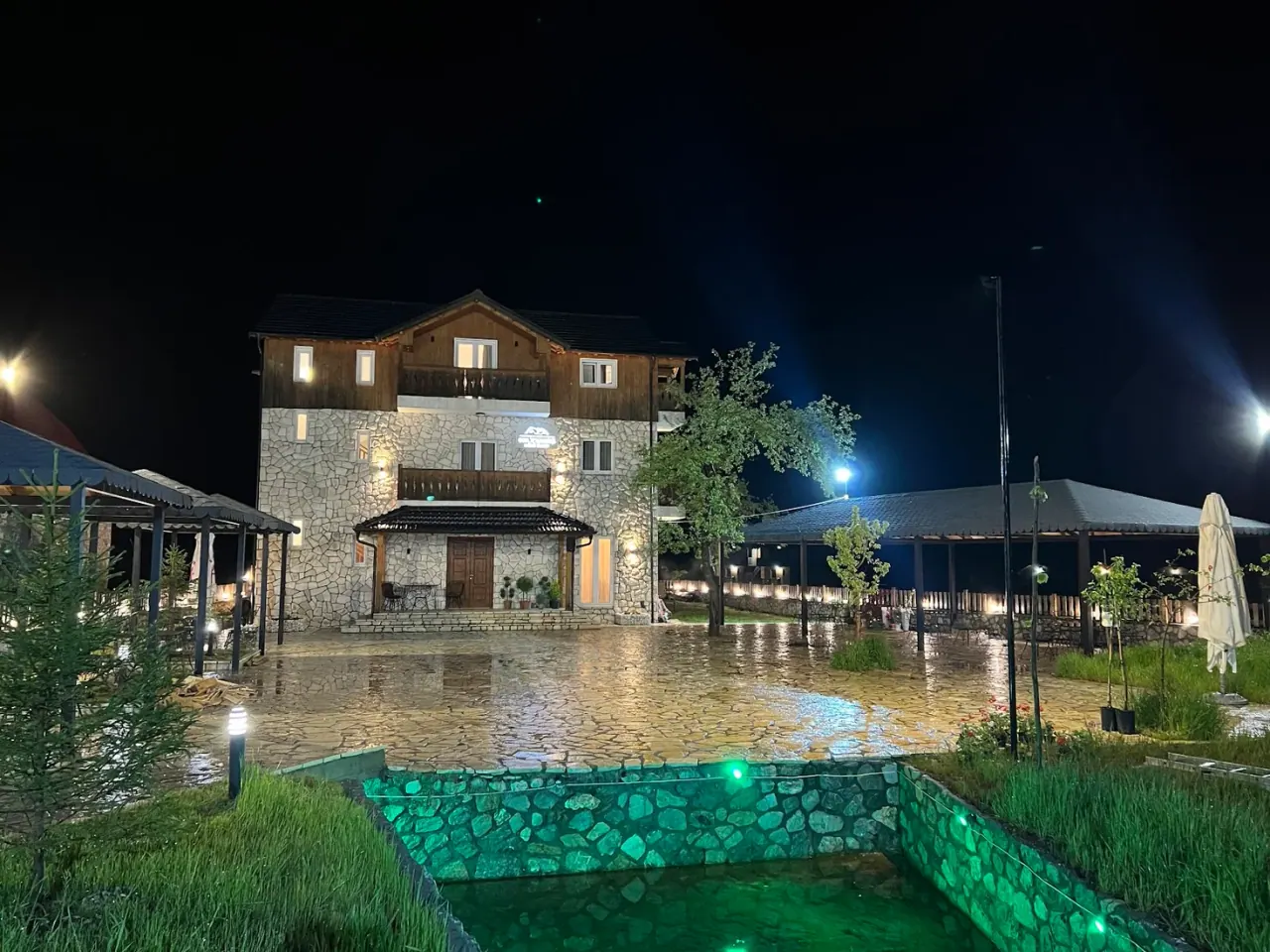 Stone-and-timber guesthouse lit at night beside a garden water feature at Oda n'Bjeshkë in Valbona