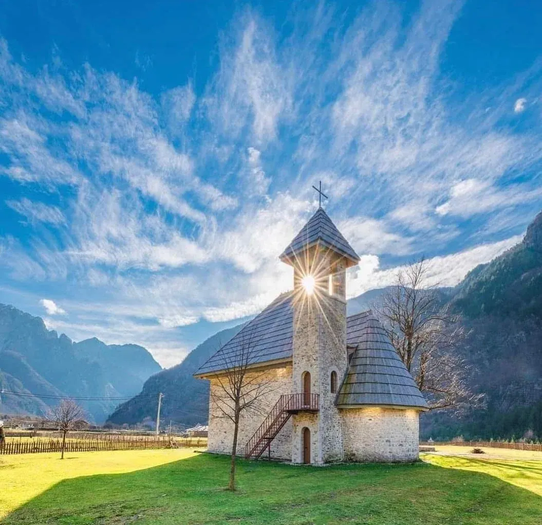 The Theth church backlit by the sun through its bell tower, with snow-dusted peaks behind