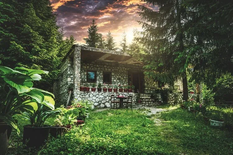 Stone guesthouse with a covered porch and pots of geraniums in a wooded garden at Guesthouse Hyrmet Demushi in the Valbona valley