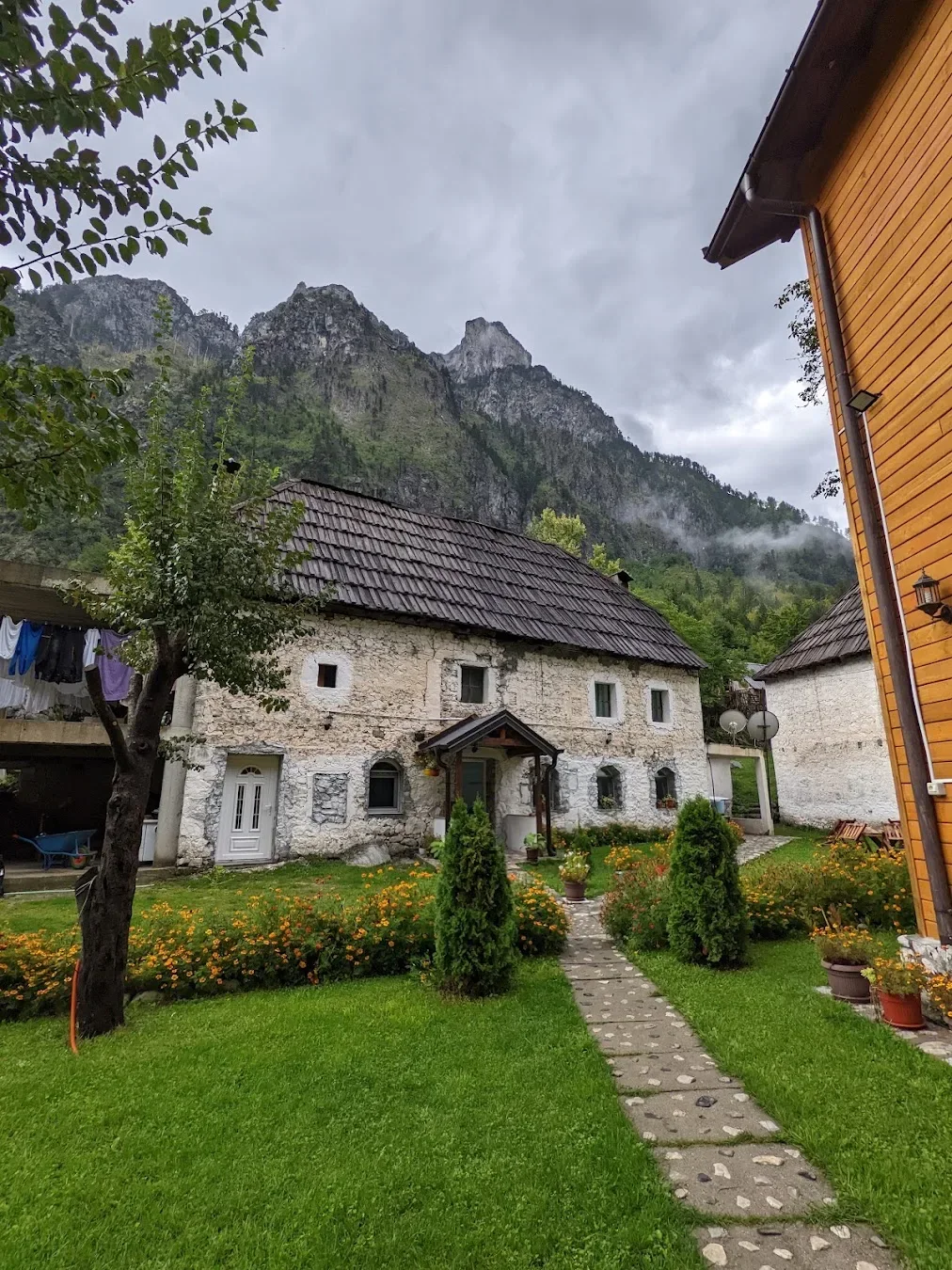Old white stone house with a shingled roof and marigold beds beside a timber building at Guesthouse Mehmeti in the Valbona valley