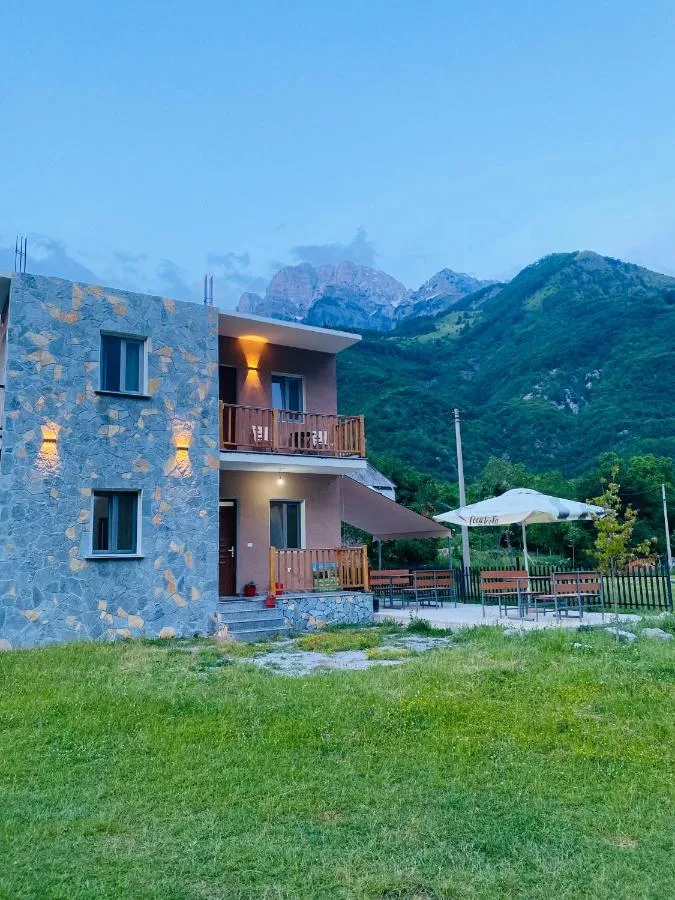 Stone-faced house with balconies and a parasol on the lawn below the peaks at Guesthouse Natyra in Theth