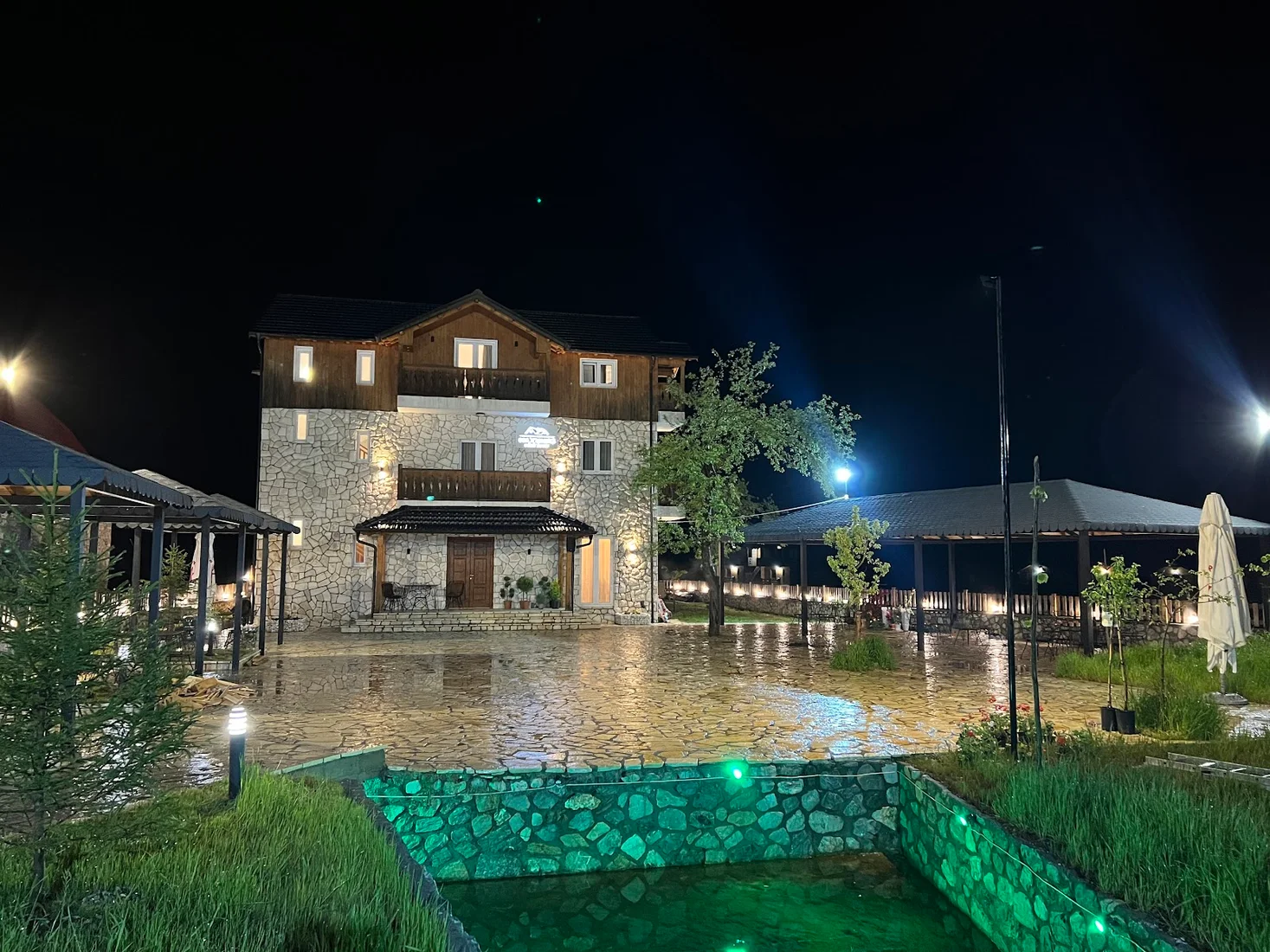 Stone-and-timber guesthouse lit at night beside a garden water feature at Oda n'Bjeshkë in Valbona