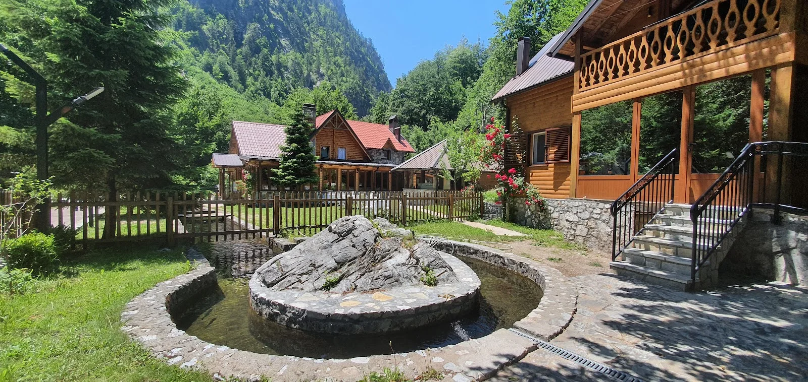 Timber and stone chalets of Guest House Rilindja with a stone fountain below forested peaks in Valbona