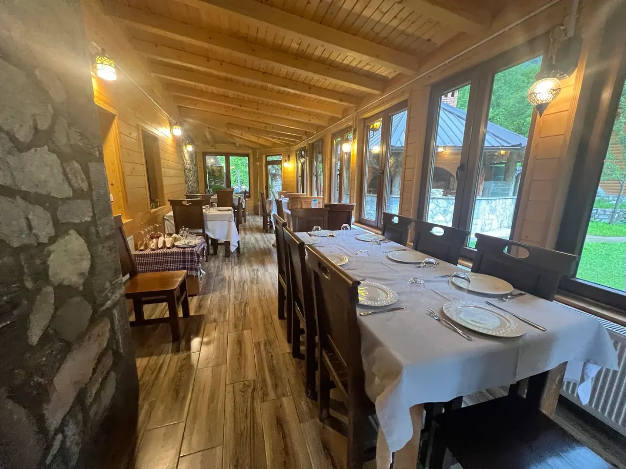Timber-and-stone dining room with a glassed veranda framing the peaks at Rilindja Restaurant in Valbonë